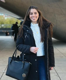 A woman stands outdoors on a plaza wearing a winter coat and smiling