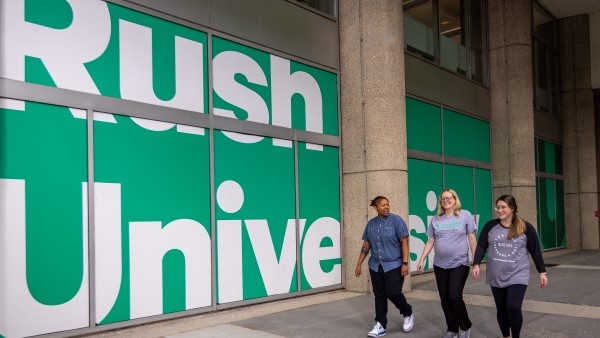 Three students walk together and smile outside in front of a set of large windows covered with a RUSH University sign.
